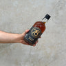 A hand holding a bottle of 78 Degrees Native Grain Whisky with a label showcasing the brand and product name, against a concrete background.