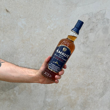 A hand holding a bottle of Amrut Cask Strength Whisky with a concrete background.