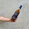 A hand holding a bottle of Amrut Cask Strength Whisky with a concrete background.