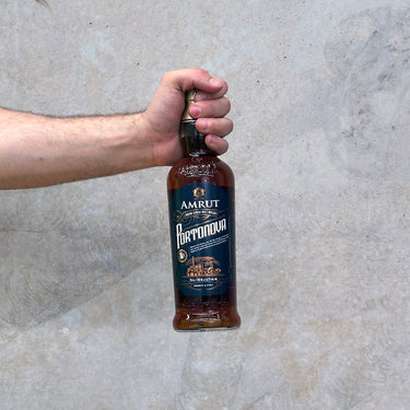A hand holding a bottle of Amrut Portonova Whisky with a concrete wall in the background.