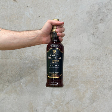 A hand holding a bottle of Amrut Spectrum 2022 Whisky with a label predominantly in dark colors, against a light concrete background.