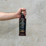 A hand holding a bottle of Amrut Spectrum 2022 Whisky with a label predominantly in dark colors, against a light concrete background.