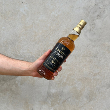 A hand holding a bottle of Amrut Triparva Triple Distilled Whisky with a clear label, in front of a grey background.