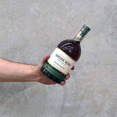 Hand holding a bottle of Archie Rose whiskey against a gray concrete background