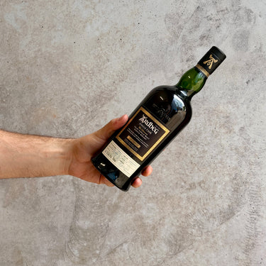 Hand holding a bottle of Ardbeg Single Cask whisky against a concrete wall.