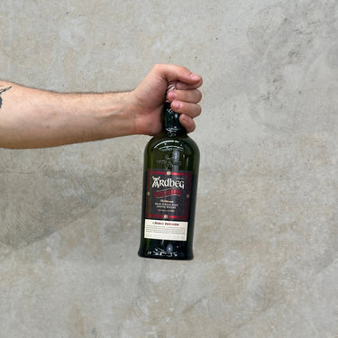A hand holding a bottle of Ardbeg Spectacular Whisky with a concrete wall in the background.
