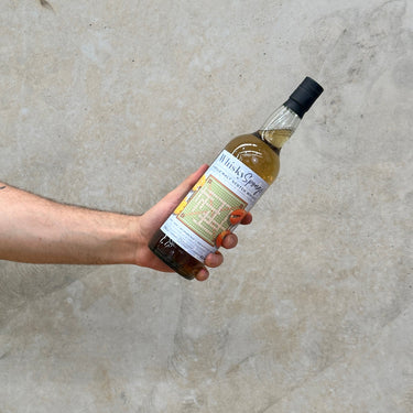 A hand holding a bottle of Ardmore WhiskySponge Edition No. 76 1997 24 Year Old against a textured grey background.