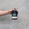 A hand holding a bottle of Backwoods Rye Shiraz Cask Finish Whisky against a concrete background.