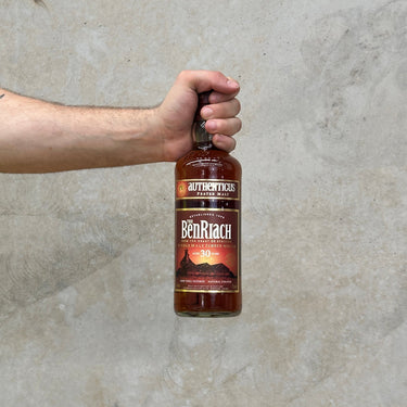 Hand holding a bottle of The Ben Riach aged 30 Years whisky against a concrete wall.