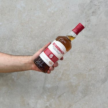 A hand holding a bottle of Benromach 15 Year Old whisky against a grey concrete background.
