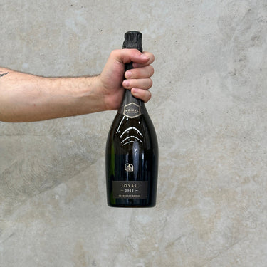 A hand holding a bottle of Boizel 2012 Joyau Champagne, which is presented in a black bottle with gold trim against a grey background. Presents in a gift box.