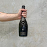 A hand holding a bottle of Boizel 2012 Joyau Champagne, which is presented in a black bottle with gold trim against a grey background. Presents in a gift box.