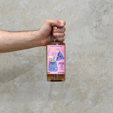 A hand holding a bottle of That Boutique-y Whisky Company Aberlour 31 Year Old whisky with a pink label, against a grey concrete background.