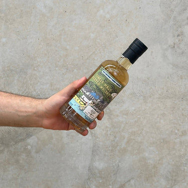 A hand holding a bottle of That Boutique-y Whisky Company Bunnahabhain 29 Year Old whisky with a label that features an image of the distillery.