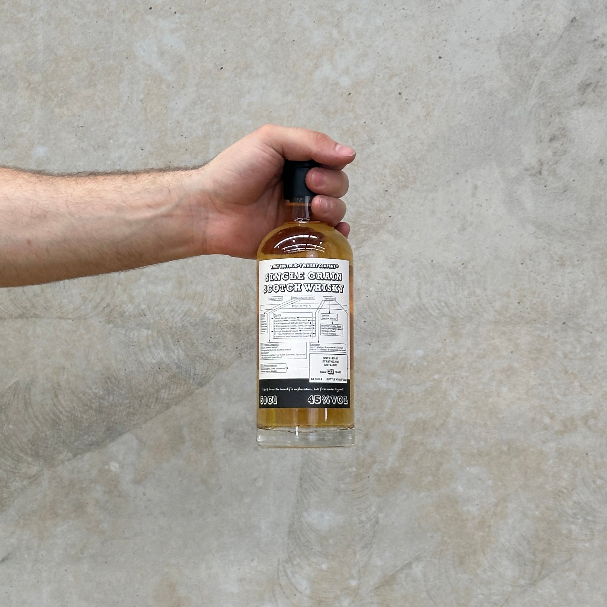 A hand holding a bottle of That Boutique-y Whisky CompanyStrathclyde 31 Year Old - Batch 4 whisky against a grey concrete background.