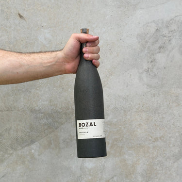 A hand holding a bottle of Bozal Castilla Ancestral Mezcal against a grey background.