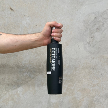 A hand holding a bottle of Bruichladdich Octomore 6.1 Whisky, which has a black label and is shown against a grey concrete background.