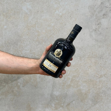 A hand holding a bottle of Bunnahabhain 25 Year Old Whisky against a concrete background.