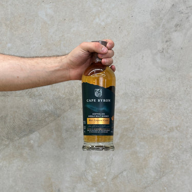 Hand holding a bottle of Cape Byron Mac. Liqueur Cask against a neutral background