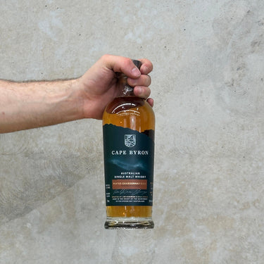 Hand holding a bottle of Cape Byron Australian Single Malt Whisky against a neutral background