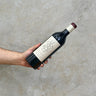 A hand holding a bottle of Chétaito Revelette Le Grand Rouge wine, with a plain concrete background.