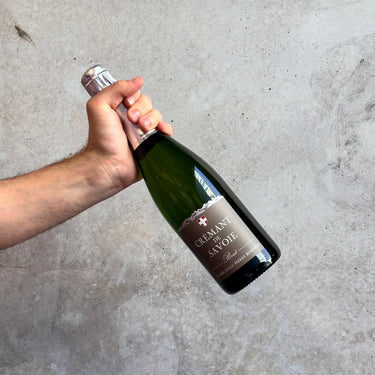 Hand holding a bottle of Cremant de Savoie Champagne Sparkling Wine against a gray concrete background