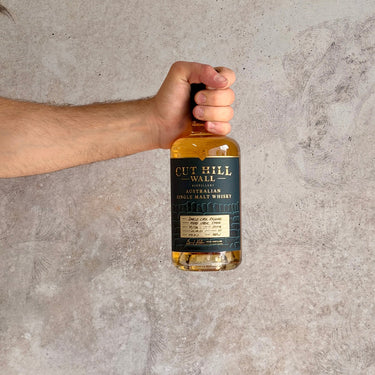 Hand holding a bottle of Cut Hill Wall Australian single malt whisky against a gray concrete background