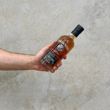 A hand holding a bottle of Balmenach Darkness 16 Year Old Oloroso Cask single malt Scotch whisky against a grey background.