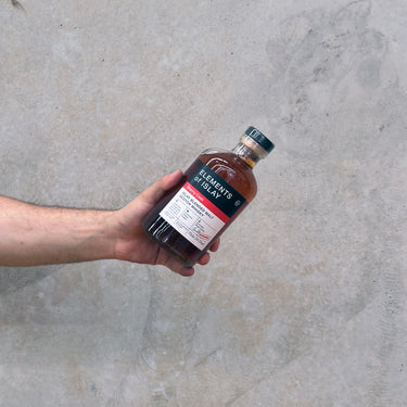 A hand holding a bottle of Elements of Islay Sherry Cask - Cask Strength Whisky against a grey concrete background.