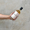 A hand holding a bottle of Arran Jean Boyer Gifted Stills Of Scotland 1996 Whisky against a grey concrete background.