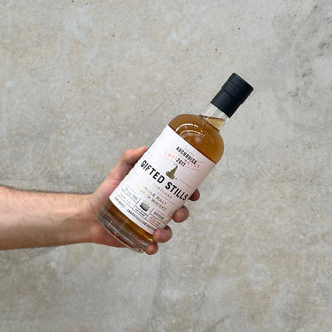 A hand holding a bottle of Auchriosk Jean Boyer Gifted Stills Of Scotland 2001 Whisky against a textured grey background.