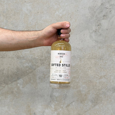 A hand holding a bottle of Benriach Jean Boyer Gifted Stills Of Scotland 2012 Whisky against a grey concrete background.