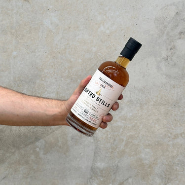A hand holding a bottle of Tullibardine Jean Boyer Gifted Stills Of Scotland 2016 Whisky with a label and cap, against a grey concrete background.