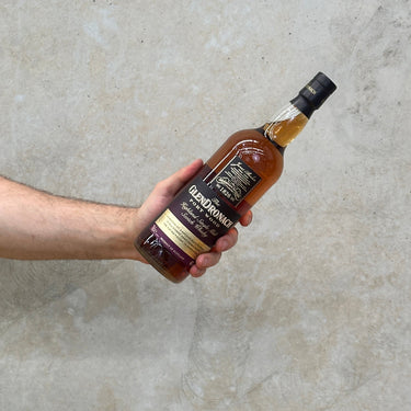 A hand holding a bottle of Glendronach Port Wood Whisky, against a grey concrete background.