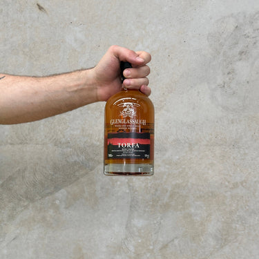 A hand holding a bottle of Glenglassaugh Torfa single malt Scotch whisky against a grey concrete background.