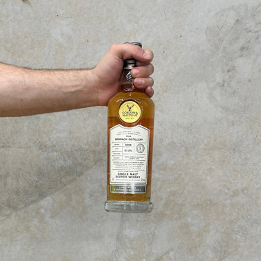 A hand holding a bottle of Gordon & Macphail Connoisseurs Choice Benriach 1999 Whisky against a grey background.