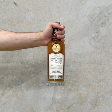 A hand holding a bottle of Gordon & Macphail Connoisseurs Choice Strathmill 1991 Whisky against a grey background.