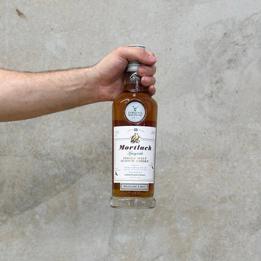 Hand holding a bottle of Gordon & Macphail Mortlach whisky against a concrete wall.