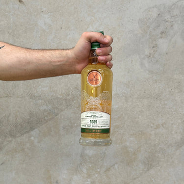 A bottle of Gordon & Macphail Discovery Tomatin 2009 Bourbon Cask Whisky held in hand, against a grey background.