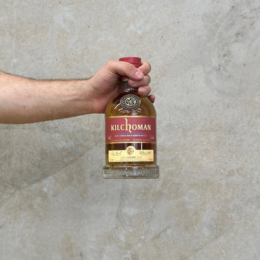 A hand holding a bottle of Kilchoman Casado Whisky with a concrete background.