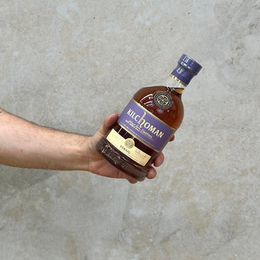 A hand holding a bottle of Kilchoman Sanaig Whisky against a grey concrete background.
