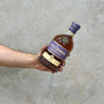 A hand holding a bottle of Kilchoman Sanaig Whisky against a grey concrete background.