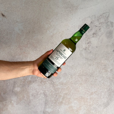 Hand holding a bottle of Laphroaig 15-year-old Islay single malt Scotch whisky against a gray background