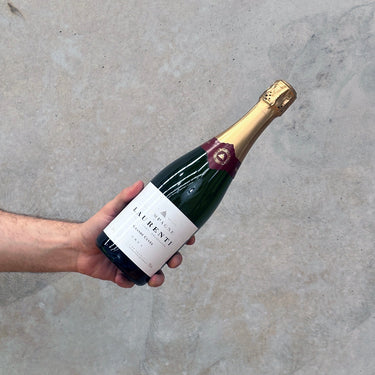 A hand holding a bottle of Laurenti NV Grande Cuvée champagne, with a concrete background.