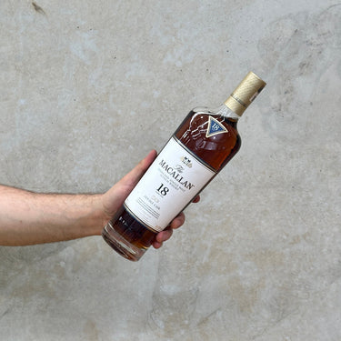 A hand holding a bottle of Macallan 18 Year Old Double Cask Whisky with a label that includes the brand name and the age statement.