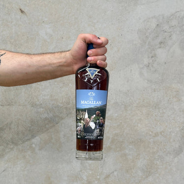 A hand holding a bottle of Macallan Sir Peter Blake Whisky with a blue label, against a grey concrete background.