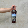 A hand holding a bottle of Macallan Sir Peter Blake Whisky with a blue label, against a grey concrete background.