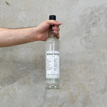 A hand holding a clear glass bottle of Mezcalosfera Madrecuixe Tobaxiche mezcal against a grey concrete background.