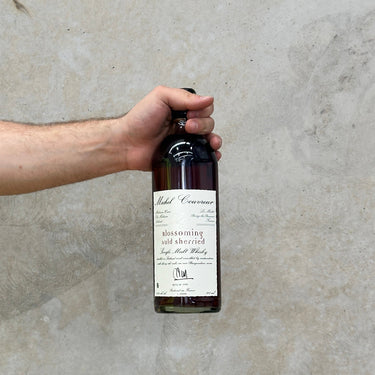 A hand holding a bottle of Michel Couvreur Blossoming Auld Sherry whisky against a grey concrete background.