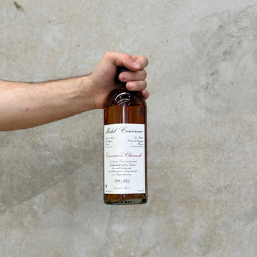 A hand holding a bottle of Michel Couvreur Clearach Whisky with a label visible, against a grey concrete background.